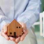 Hands gently holding a small wooden model house, symbolizing home ownership.