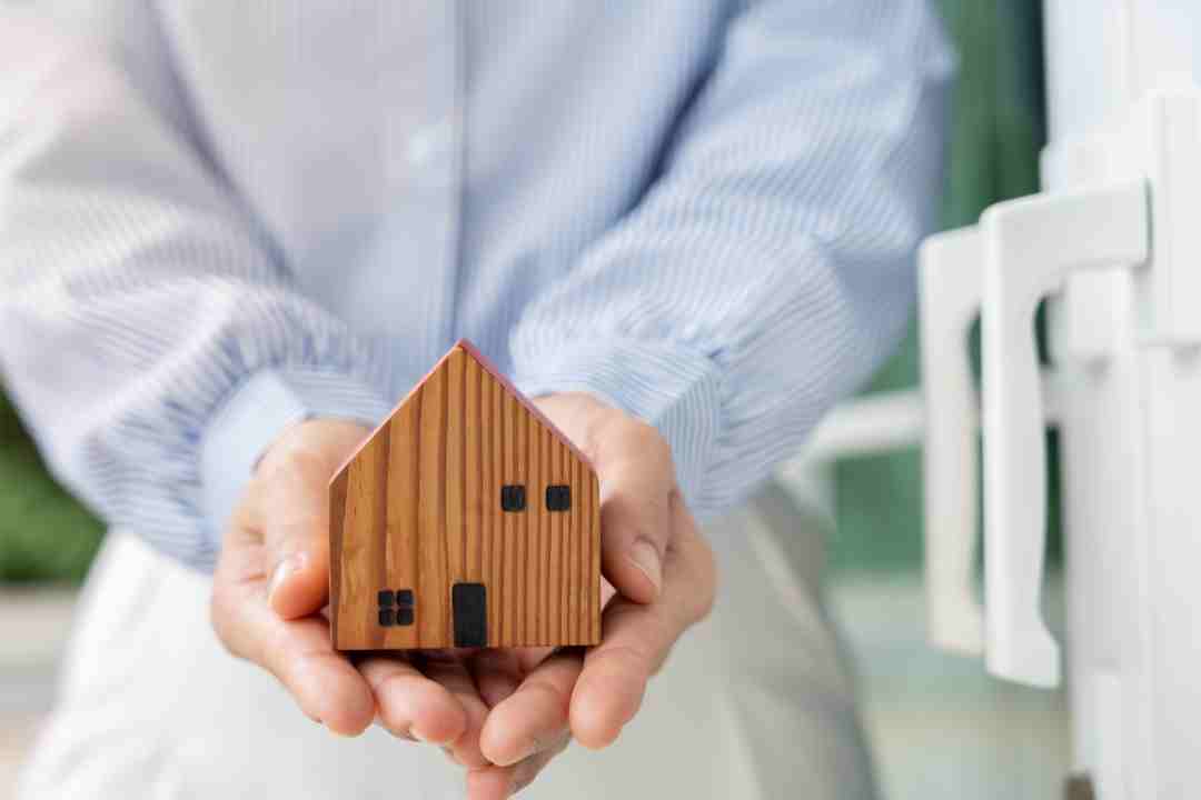 Hands gently holding a small wooden model house, symbolizing home ownership.